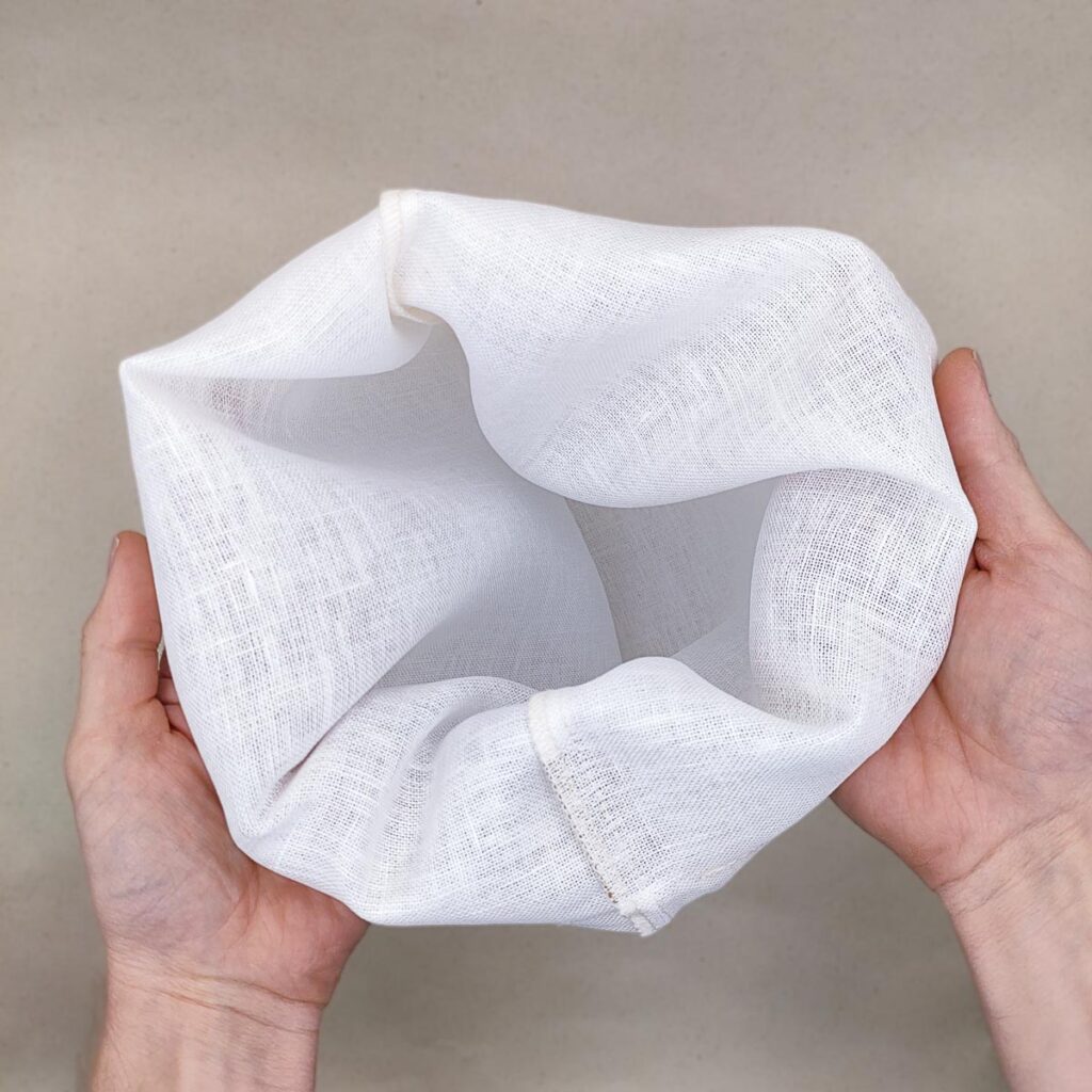 2 Hände öffnen einen weissen Leinenfilterbeutel sodass man in das leere innere blicken kann.